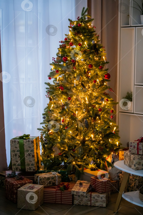 Скачать Украшенная рождественская елка со светящимися огнями и подарочной коробкой под ней в гостиной. фотосток Ozero