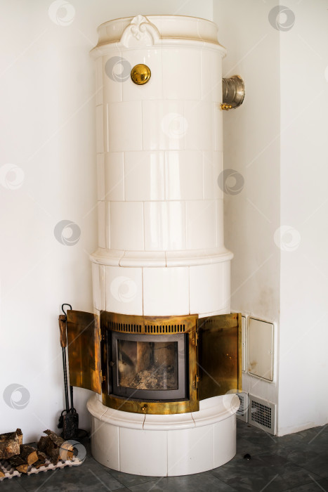 Скачать Белая изразцовая печь с открытыми латунными дверцами и дровами для растопки рядом с ней. Традиционное внутреннее отопительное устройство для дома фотосток Ozero