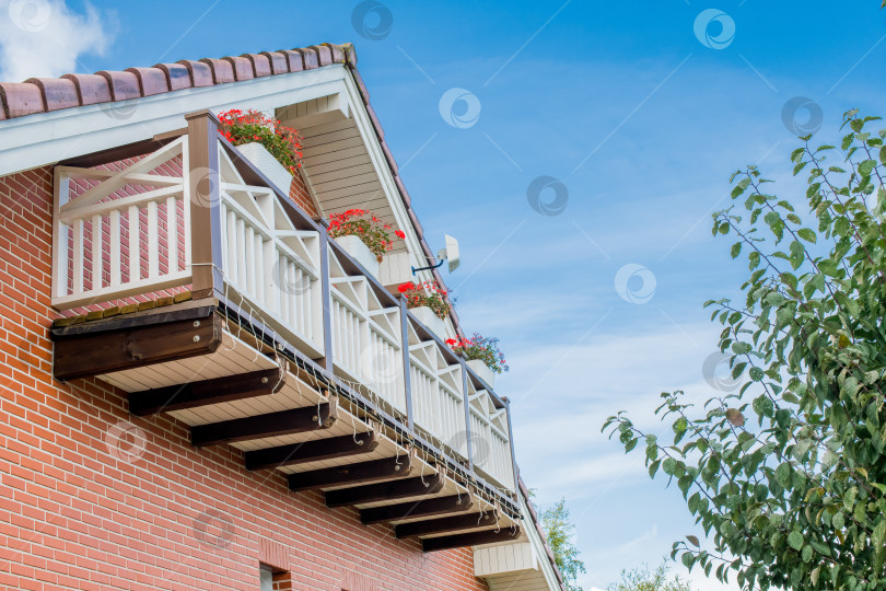 Скачать Балкон дома, украшенный красными цветами на фоне ясного голубого неба, демонстрирует архитектурные детали и природную красоту фотосток Ozero