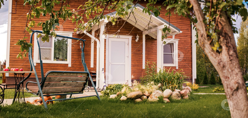 Скачать Уютный деревянный дом с верандой и качелями во дворе. Концепция летней загородной жизни и отдыха на свежем воздухе. фотосток Ozero