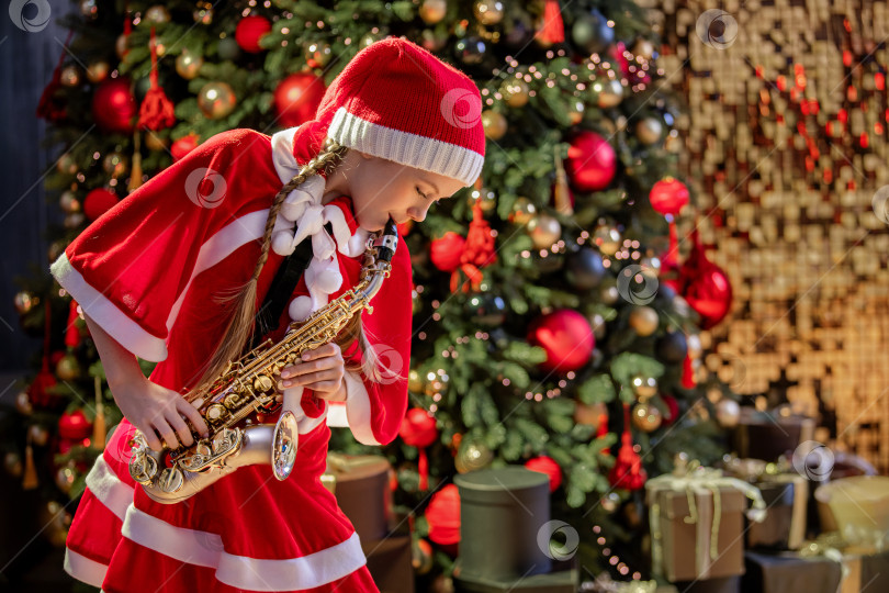 Скачать Девочка в красной рождественской шапочке Санты играет на саксофоне возле рождественской елки в украшенном зале. фотосток Ozero