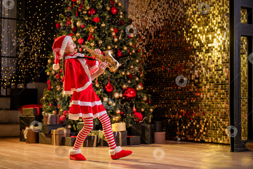 Скачать Девочка в красной рождественской шапочке Санты играет на саксофоне возле рождественской елки в украшенном зале. фотосток Ozero