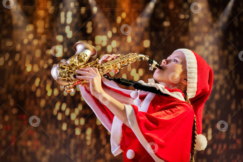 Скачать Девочка в красной рождественской шапочке Санты играет на саксофоне в украшенном зале. фотосток Ozero