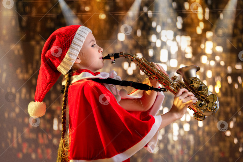 Скачать Девочка в красной рождественской шапочке Санты играет на саксофоне в украшенном зале. фотосток Ozero