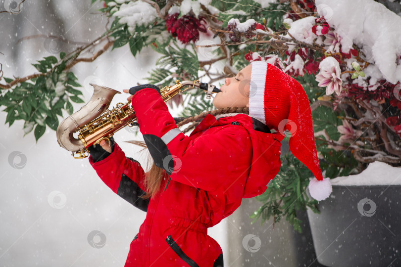 Скачать Маленькая девочка в красной рождественской шапочке Санты играет на саксофоне во время снегопада на городской улице. фотосток Ozero
