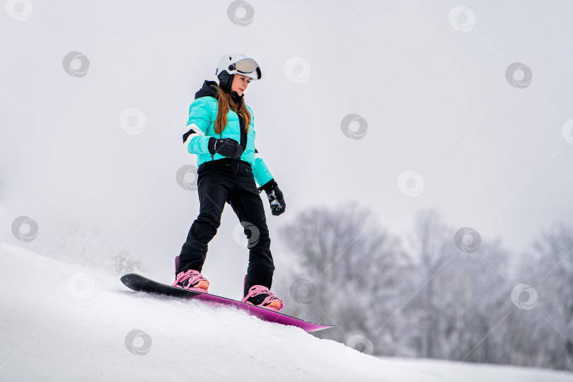 Скачать Молодая сноубордистка, скользящая по заснеженному склону в стильном зимнем снаряжении. Захватывающие зимние виды спорта. фотосток Ozero