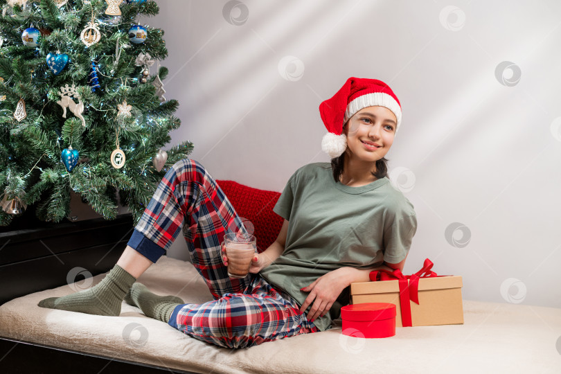 Скачать женщина в шапке Санты и пижаме держит горячий шоколад рядом с рождественским столом. фотосток Ozero