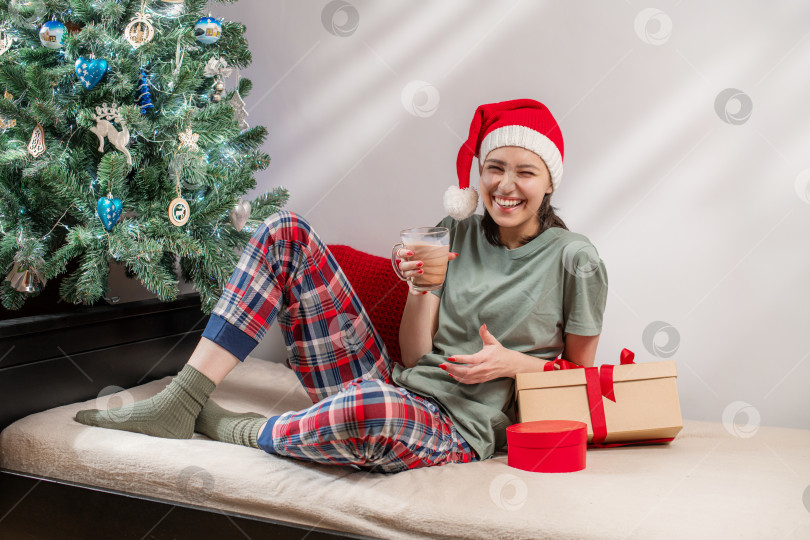 Скачать женщина в шапке Санты и пижаме держит горячий шоколад рядом с рождественским столом. фотосток Ozero