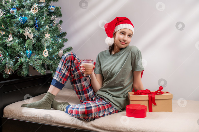Скачать женщина в шапке Санты и пижаме держит горячий шоколад рядом с рождественским столом. фотосток Ozero