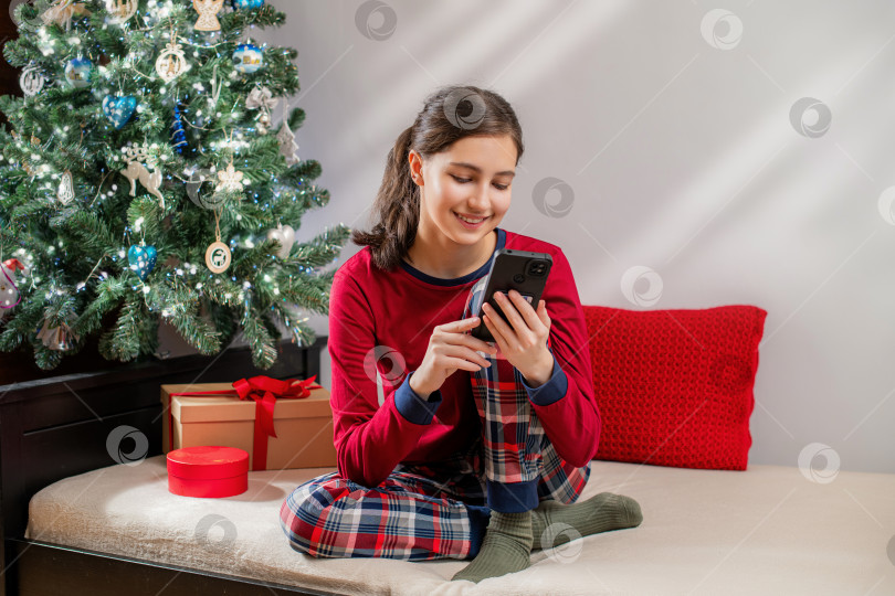 Скачать Улыбающаяся девушка в красной пижаме пользуется мобильным телефоном возле рождественской елки фотосток Ozero