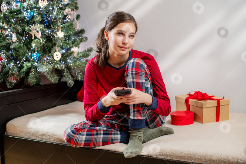 Скачать Улыбающаяся девушка в красной пижаме пользуется мобильным телефоном возле рождественской елки фотосток Ozero