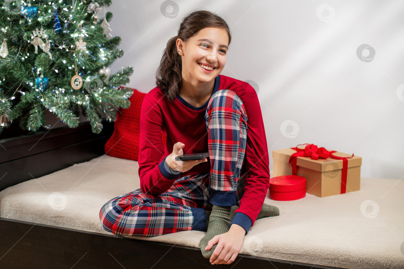 Скачать Улыбающаяся девушка в красной пижаме пользуется мобильным телефоном возле рождественской елки фотосток Ozero
