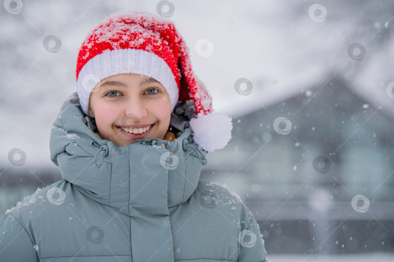 Скачать Молодая девушка-модель со счастливой улыбкой, одетая в красную шапку Санты во время зимнего снегопада фотосток Ozero