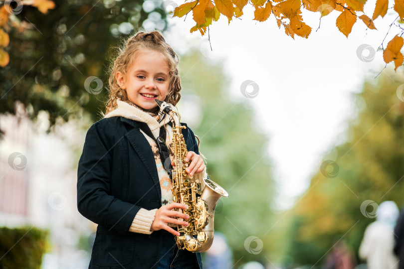 Скачать Счастливая маленькая девочка осенью стоит на улице с саксофоном в руках. фотосток Ozero