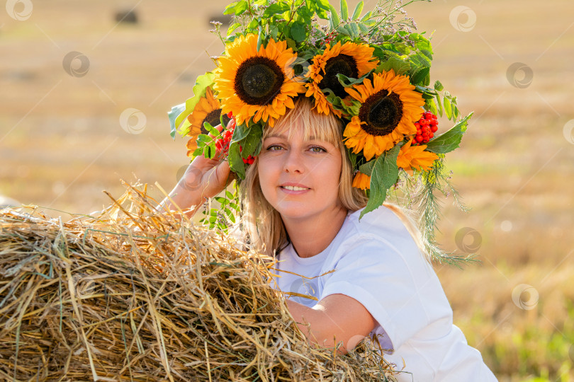 Скачать женщина в ярком венке из подсолнухов, прислонившаяся к стогу сена на залитом солнцем поле. Праздник урожая. фотосток Ozero
