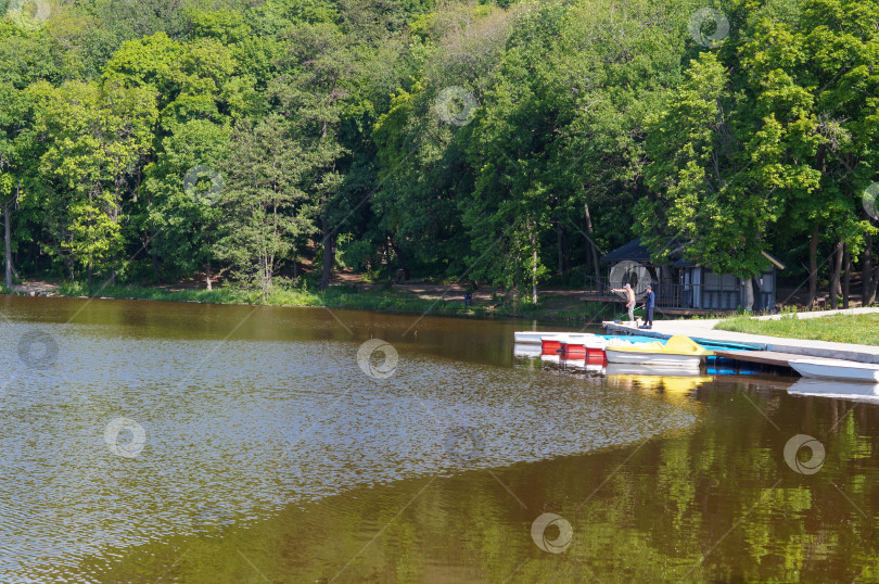Скачать Саратов, Россия - 25 мая 2025 года.  Густой лес окружает озеро с причалом, где рыбаки ловят рыбу фотосток Ozero