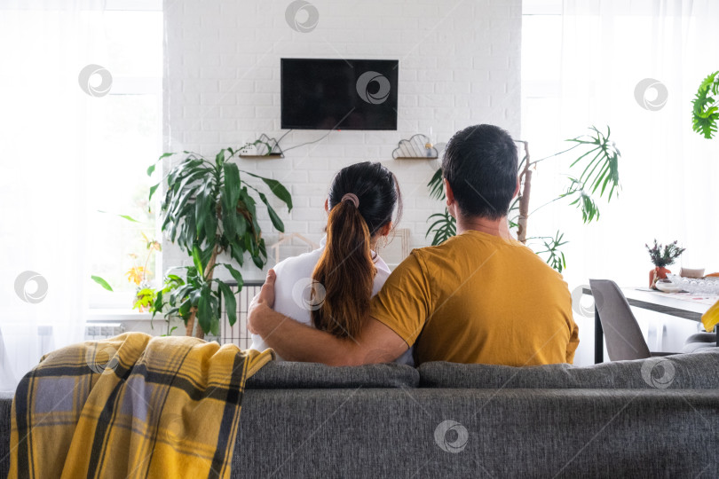 Скачать Мужчина и женщина дома в белом интерьере сидят на диване и смотрят телевизор, макет черного экрана. Домашний отдых, досуг, любовь, семья, свидание фотосток Ozero
