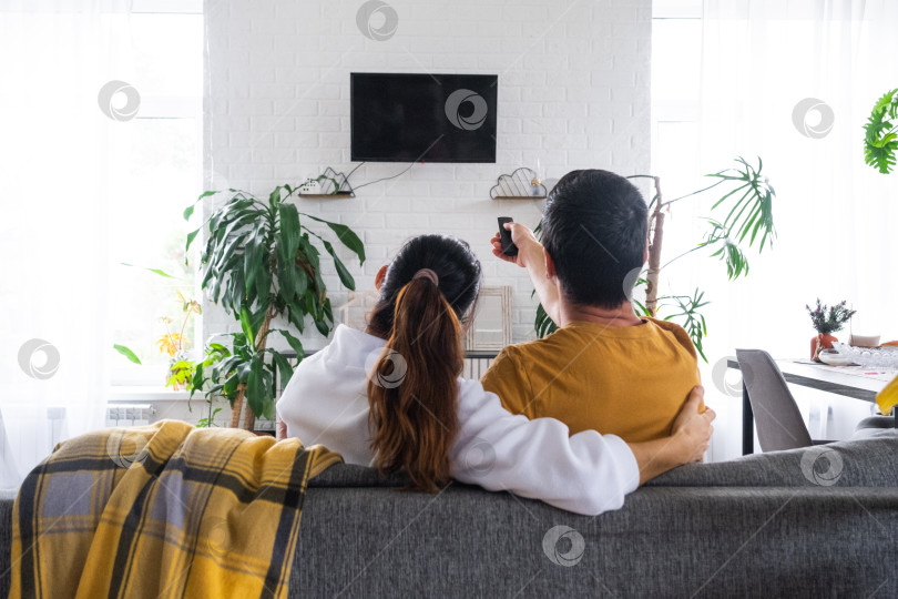 Скачать Мужчина и женщина дома в белом интерьере сидят на диване и смотрят телевизор, макет черного экрана. Домашний отдых, досуг, любовь, семья, свидание фотосток Ozero