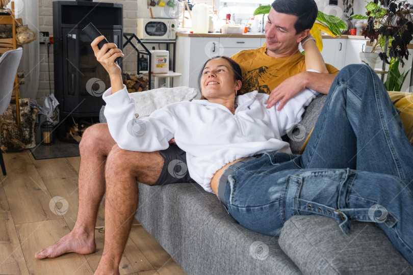 Скачать Мужчина и женщина дома в белом интерьере сидят на диване и смотрят телевизор, макет черного экрана. Домашний отдых, досуг, любовь, семья, свидание фотосток Ozero