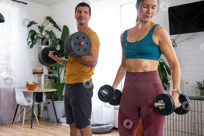 Скачать Женщина и мужчина в спортивной форме занимаются фитнесом и силовыми тренировками со штангой и гантелями дома в интерьере и кат фотосток Ozero