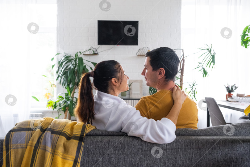 Скачать Мужчина и женщина дома в белом интерьере сидят на диване и смотрят телевизор, макет черного экрана. Домашний отдых, досуг, любовь, семья, свидание фотосток Ozero