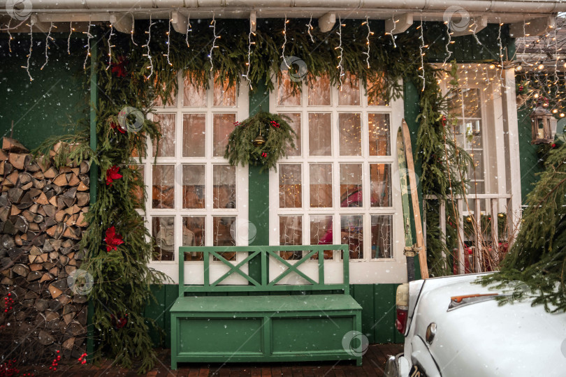 Скачать Украшенный вход в дом с зеленой скамейкой перед большим окном, украшенный праздничными рождественскими гирляндами и огнями. фотосток Ozero