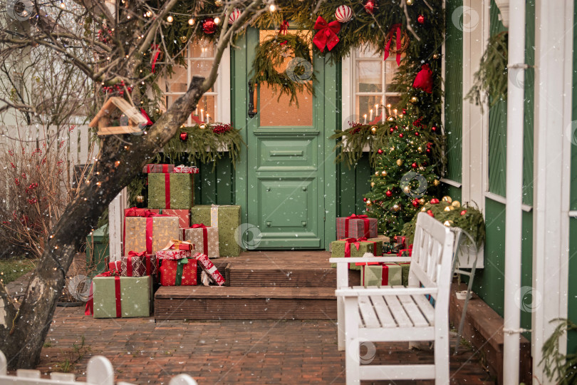 Скачать Рождественски украшенный фасад дома с дверью, подарками, вечнозеленым венком и праздничными гирляндами, на который падает снег для праздничной зимы фотосток Ozero