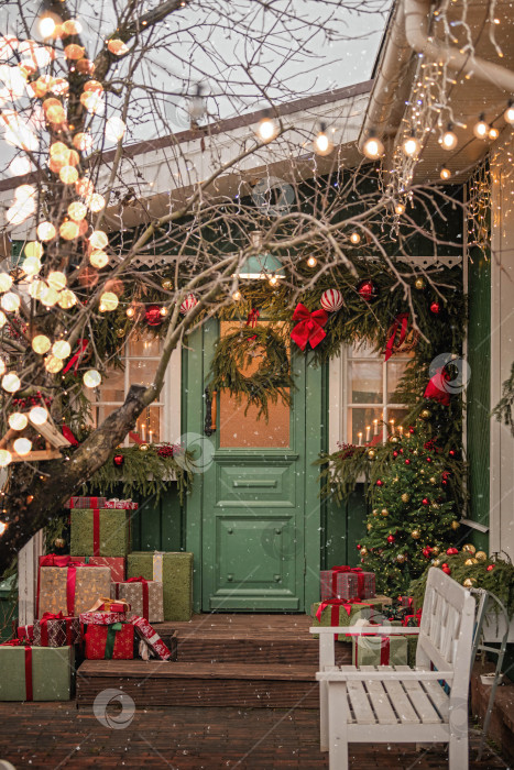 Скачать Рождественски украшенный фасад дома с дверью, подарками, вечнозеленым венком и праздничными гирляндами, на который падает снег для праздничной зимы фотосток Ozero