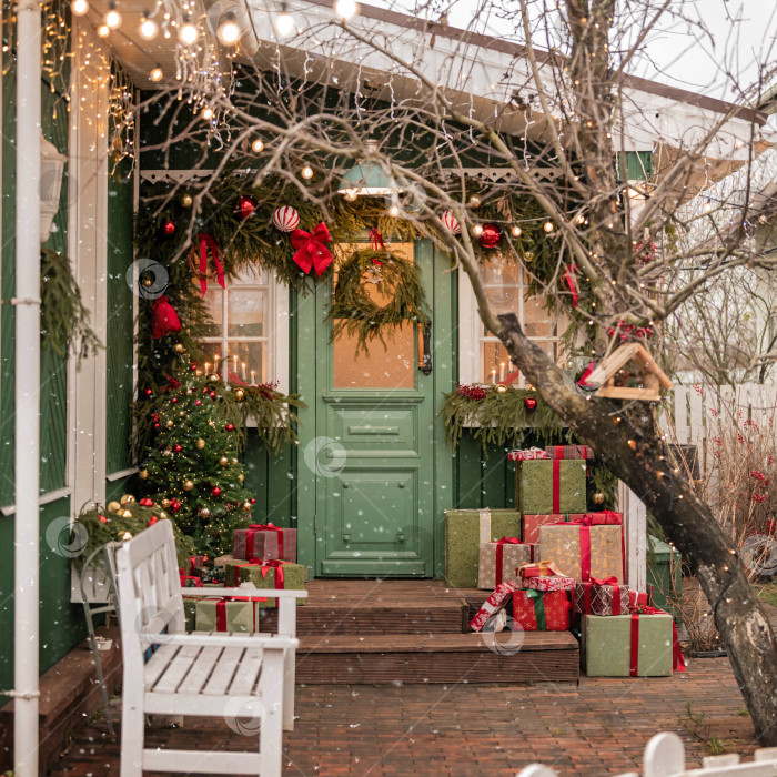 Скачать Рождественски украшенный фасад дома с дверью, подарками, вечнозеленым венком и праздничными гирляндами, на который падает снег для праздничной зимы фотосток Ozero