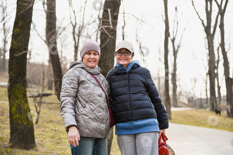 Скачать Две русские женщины 50 и 60 лет улыбаются в теплой одежде в тихом парке, демонстрируя зрелость, отсутствие морщин, умиротворение, связь и осознанный осенне-весенний момент. фотосток Ozero