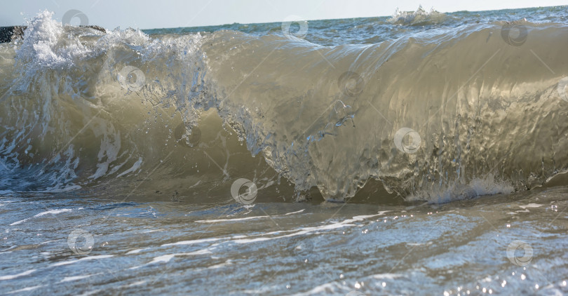 Скачать Большая волна, разбивающаяся о берег. Вода пенится и блестит на солнце. Видны брызги и пена. фотосток Ozero