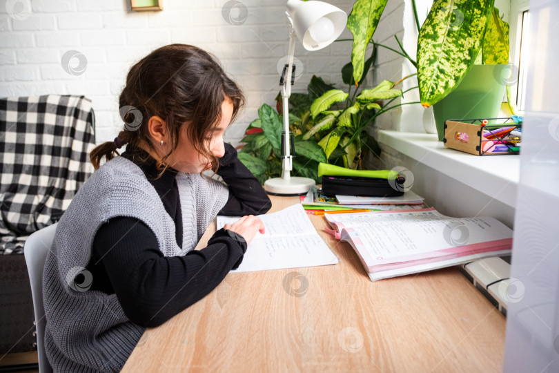 Скачать Девочка-школьница делает домашнее задание из школы за столом, что-то записывая в тетрадь фотосток Ozero