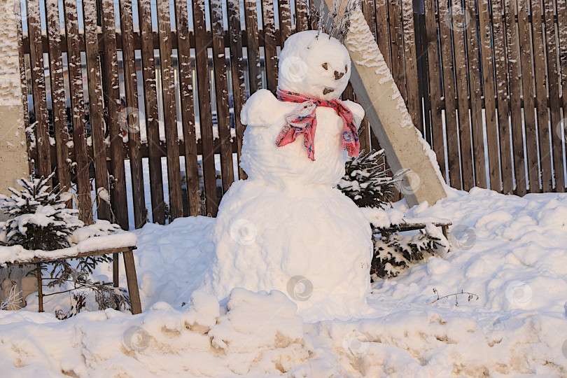 Скачать Забавный снеговик в деревне у забора с красным платочком.Зимний пейзаж Центральной России.Красивая зимняя пригласительная открытка для туристов и традиционные зимние забавы. фотосток Ozero