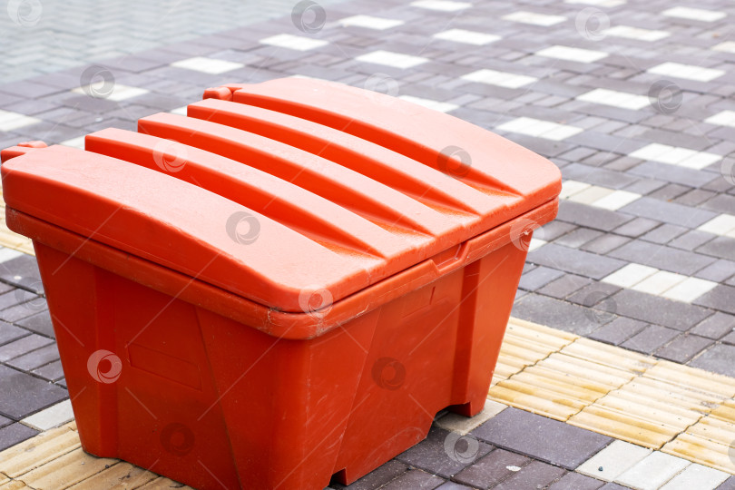 Скачать Ярко-оранжевый контейнер для песка, установленный на тротуаре рядом с тактильным ориентиром в зимних городских условиях фотосток Ozero