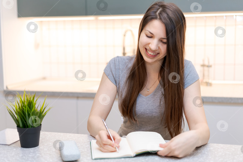 Скачать Женщина сидит за столом с блокнотом и ручкой и, улыбаясь, что-то пишет фотосток Ozero