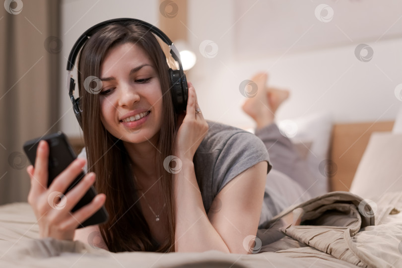 Скачать Женщина лежит на кровати с телефоном в руке и наушниками на ушах фотосток Ozero