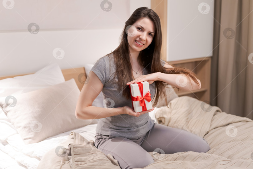 Скачать Женщина сидит на кровати, держа в руках белую подарочную коробку с красным бантом фотосток Ozero