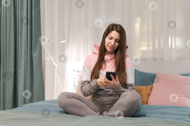 Скачать Женщина сидит на кровати и смотрит в свой мобильный телефон фотосток Ozero