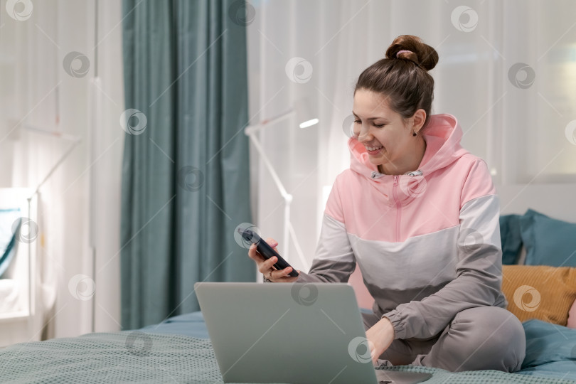 Скачать Женщина сидит на кровати с ноутбуком и сотовым телефоном в руках фотосток Ozero