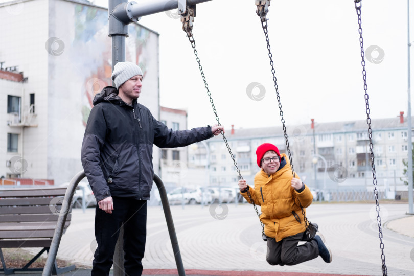 Скачать 40-летний мужчина в теплой куртке и шапке качает своего сына в желтой куртке на качелях в парке. Счастливый мальчик со своим папой на качелях. фотосток Ozero