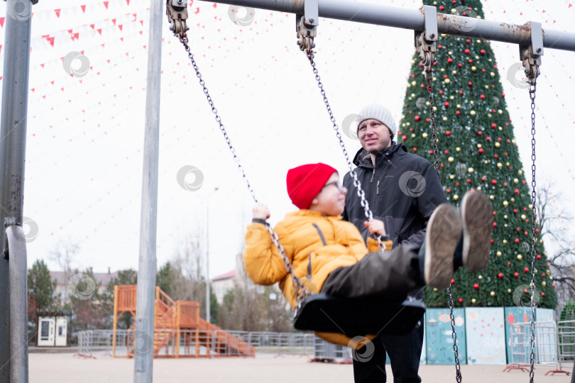 Скачать 40-летний мужчина в теплой куртке и шапке качает своего сына в желтой куртке на качелях в парке на фоне рождественской елки. фотосток Ozero