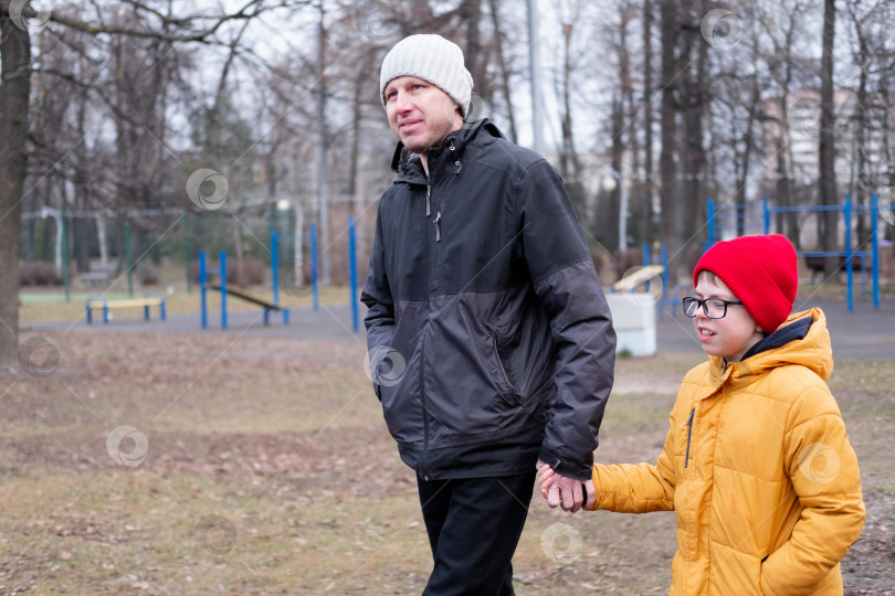 Скачать 40-летний мужчина в теплой куртке и шапке держит за руку своего сына в желтой куртке во время прогулки по парку. фотосток Ozero