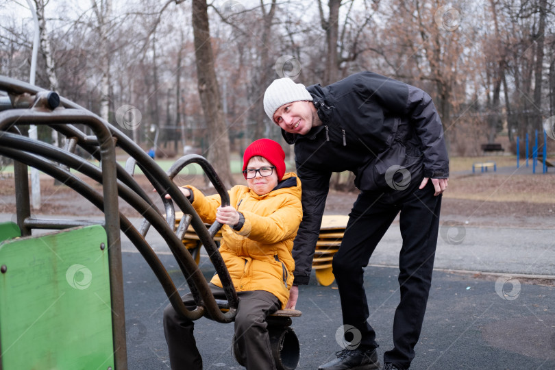 Скачать 40-летний мужчина в теплой куртке и шапке качает своего сына, одетого в желтую куртку, на металлических качелях в парке. Счастливый мальчик и его отец на качелях. фотосток Ozero