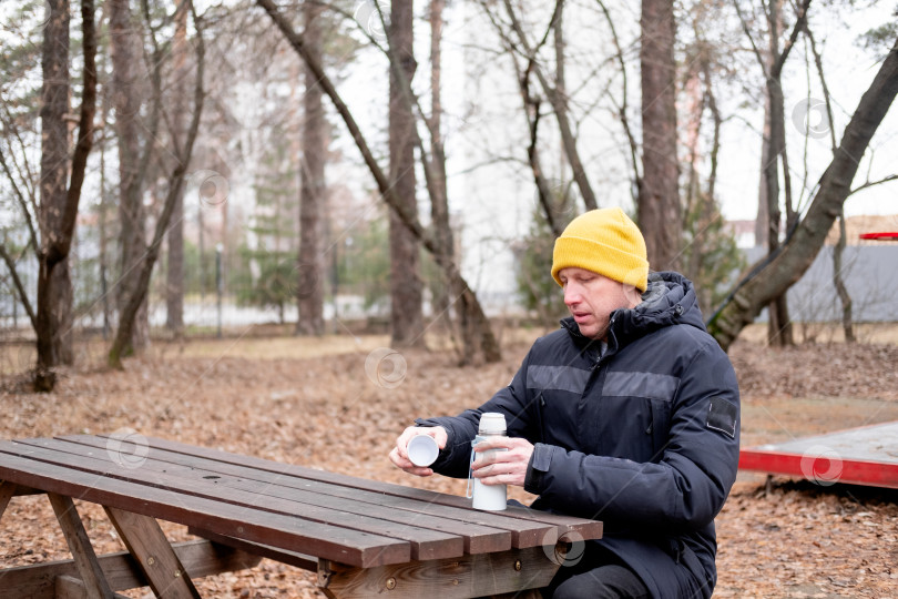 Скачать 40-летний мужчина в теплой куртке и желтой шляпе наливает горячий кофе из термоса, сидя за столиком в парке фотосток Ozero