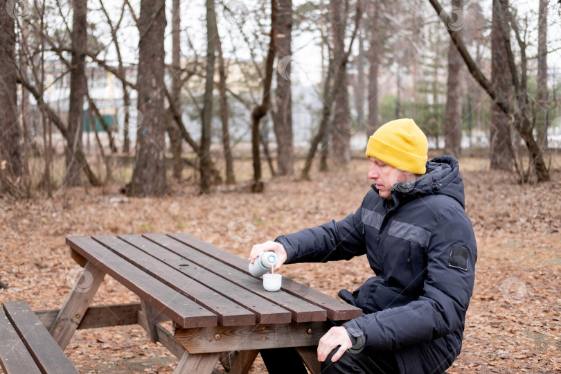 Скачать 40-летний мужчина в теплой куртке и желтой шляпе наливает горячий кофе из термоса, сидя за столиком в парке фотосток Ozero