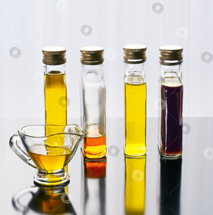 Скачать Различные растительные масла в одинаковых бутылочках расположены на темной отражающей поверхности. Перед бутылочками стоит соусник, в который налито масло. фотосток Ozero