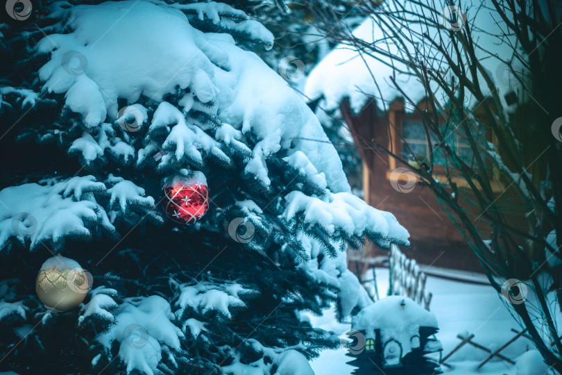 Скачать Заснеженные еловые ветки, украшенные праздничными красными и золотыми рождественскими украшениями, создают уютную праздничную атмосферу с деревенским деревянным домиком и миниатюрной зимней сценой на заднем плане фотосток Ozero