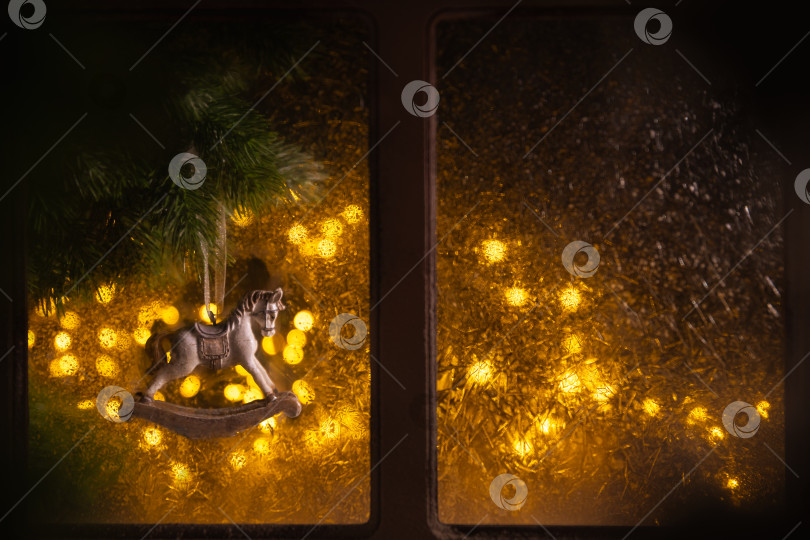 Скачать Маленькое украшение в виде лошадки-качалки, висящее на ветке рождественской елки и подсвеченное теплыми желтыми гирляндами, создает праздничную, уютную и ностальгическую атмосферу праздника за оконным стеклом фотосток Ozero