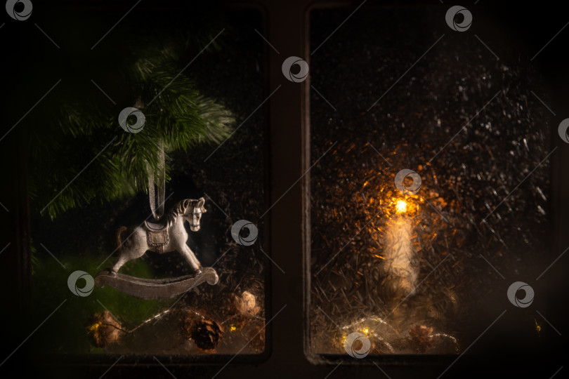 Скачать Рождественское украшение в виде лошадки-качалки, висящее на сосновой ветке с декоративными лампочками, создает уютную праздничную атмосферу, видимую через оконное стекло, а снаружи видна горящая свеча. фотосток Ozero