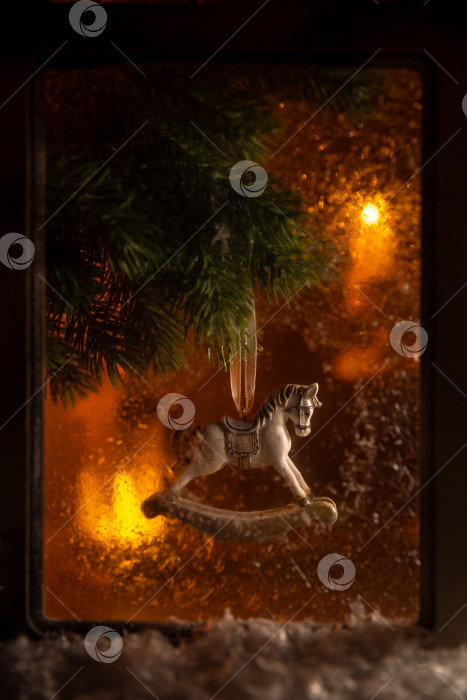 Скачать Украшение в виде лошадки-качалки свисает с ветки рождественской елки, создавая уютную праздничную атмосферу с теплыми фоновыми огнями и ностальгическим ощущением праздника фотосток Ozero